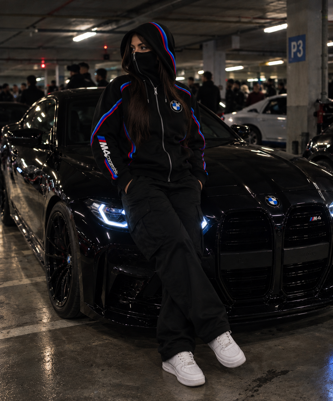 BMW design hoodie female lifestyle realistic underground car meet with integrated balaclava beside black BMW M4 Competition