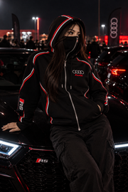 Audi RS hoodie female lifestyle — nighttime car meet, black hoodie with red and white piping, Audi chest logo, RS sleeve emblem and integrated balaclava beside black Audi RS