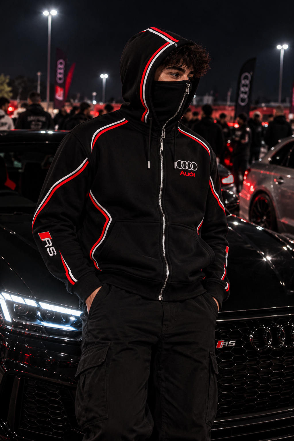 Audi RS hoodie male lifestyle — nighttime car meet, black red and white single-zip balaclava hoodie, silver Audi rings red Audi chest lettering and right sleeve RS emblem