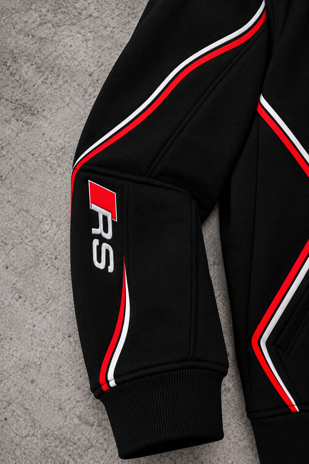Audi RS hoodie right sleeve close-up — vertical RS emblem with red square, red and white piping, black cuff, grey concrete background