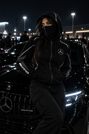 REVLINE Mercedes AMG female lifestyle at nighttime car meet, black zip balaclava hoodie, Mercedes chest emblem and AMG sleeve